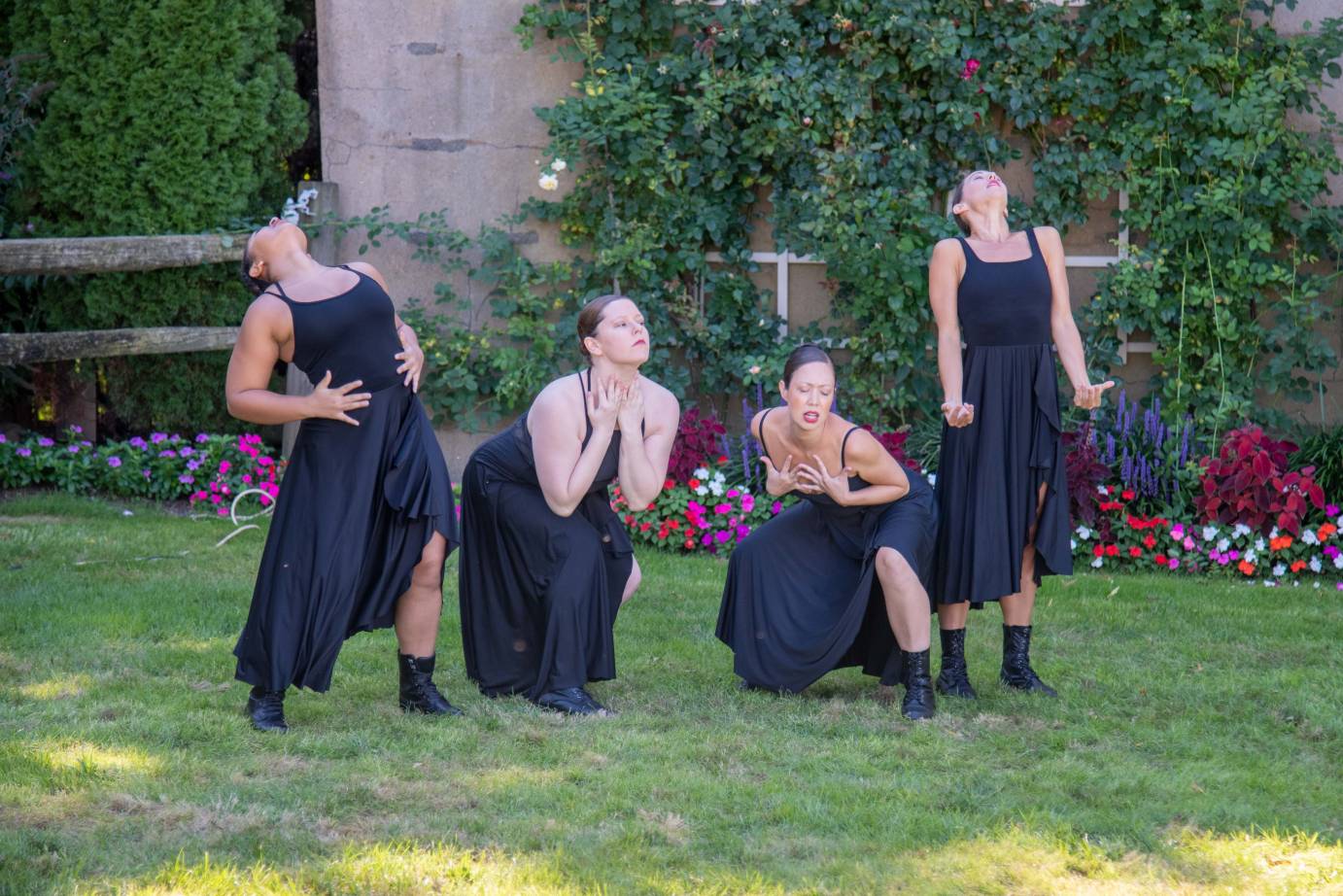 a quartet of women wearing tight black leotards with spagetti straps, black flowing skirst, and black short boots clench fists or clutch at themselves as if experience deep pain. The are in a beautiful garden setting on a green lawn with colorful flowers on the ground and a trellis with roses behind them